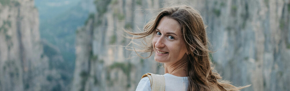 Frau mit langen Haaren, die ins Bild lächelt; im Hintergrund sind steile, felsige Klippen und Natur zu sehen.
