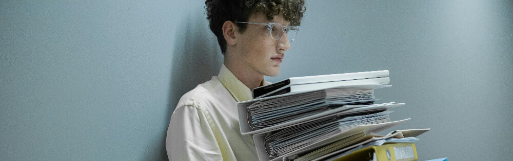 Ein junger Mann mit Locken und Brille steht an einer grauen Wand und hält einen großen Stapel Dokumente und Ordner in den Armen.