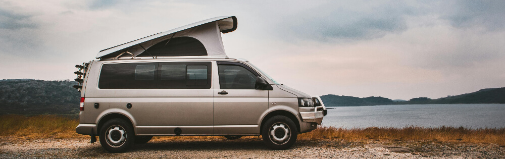 Silberner Campervan mit hochgeklapptem Dach steht an einem Seeufer mit bewölktem Himmel und Hügeln im Hintergrund.